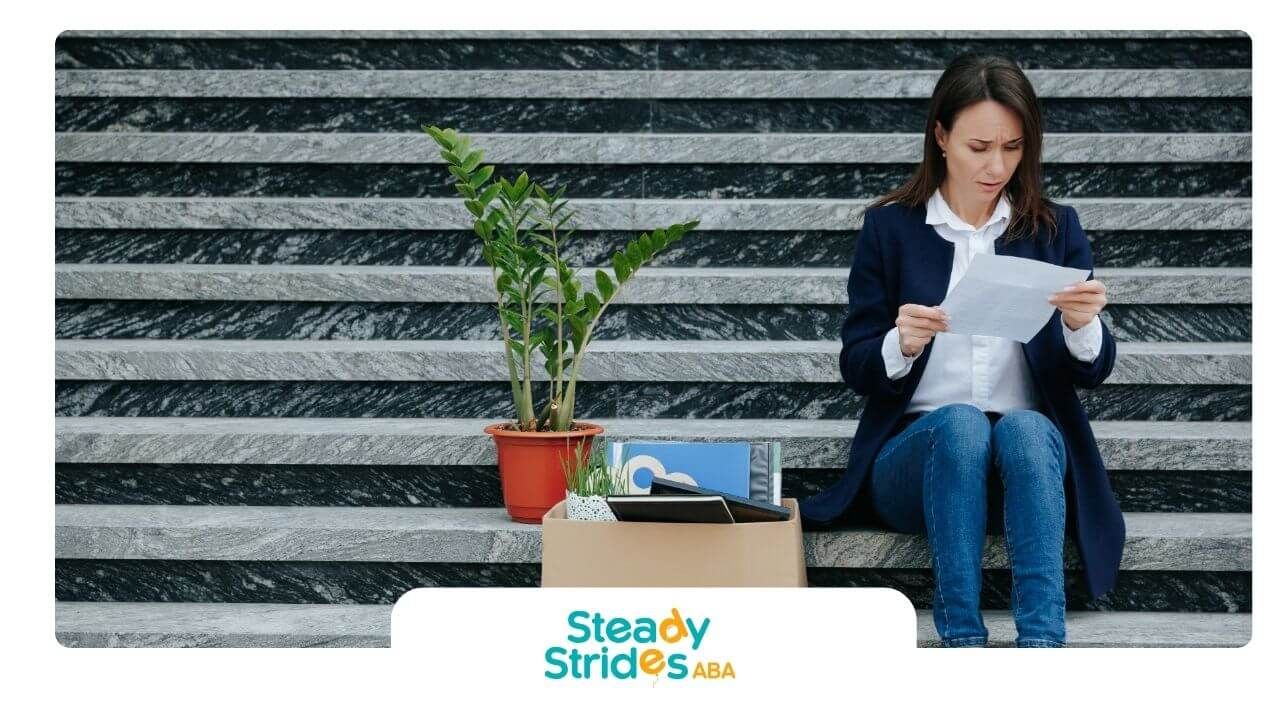 A therapist sitting on marble steps holding paperwork beside a box of office items after transition.