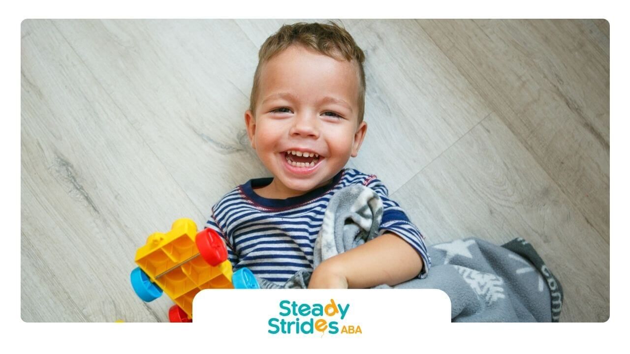 A toddler lying on the floor laughing and holding a toy car.