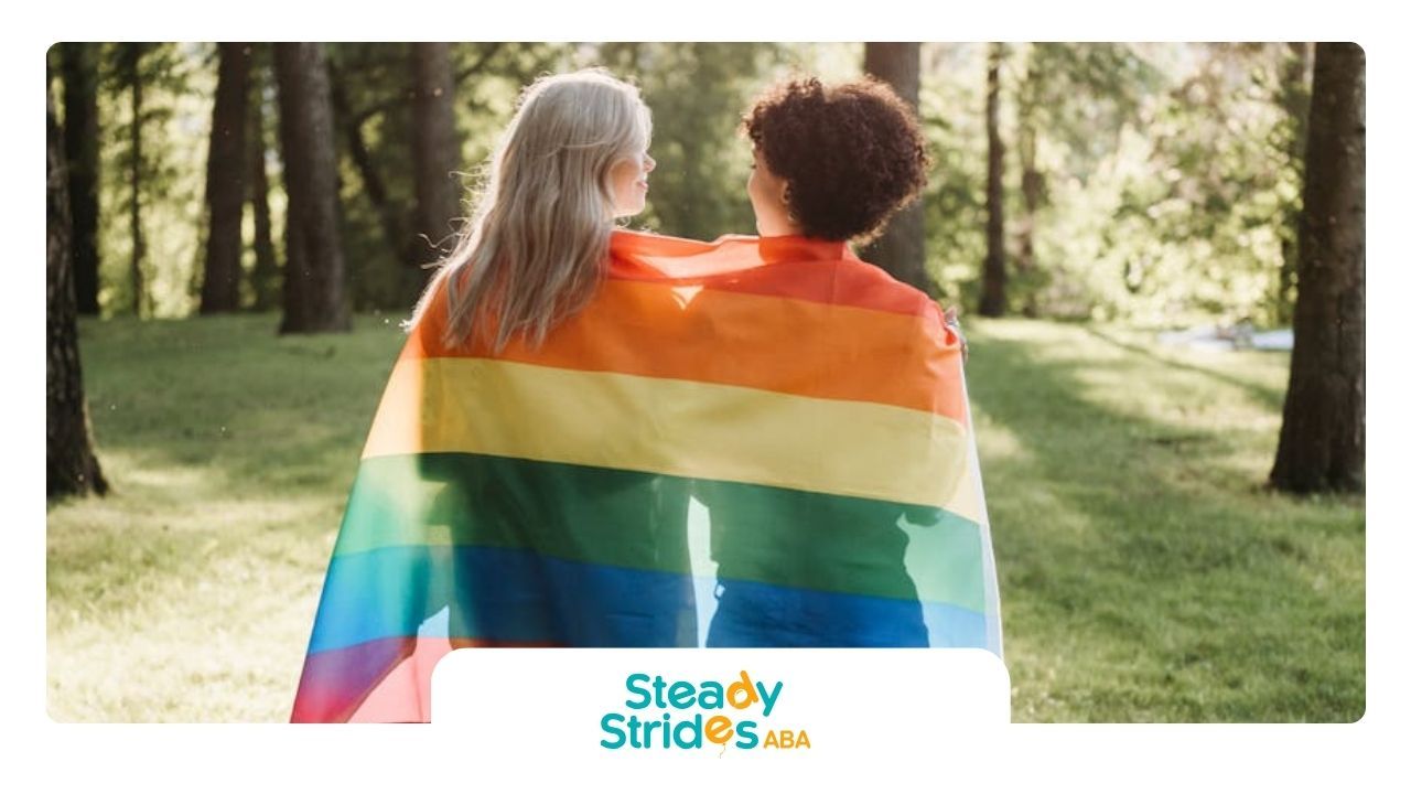 Two autistic people walking at a park in Texas, draped in a rainbow flag, embracing LGBTQ+ pride.