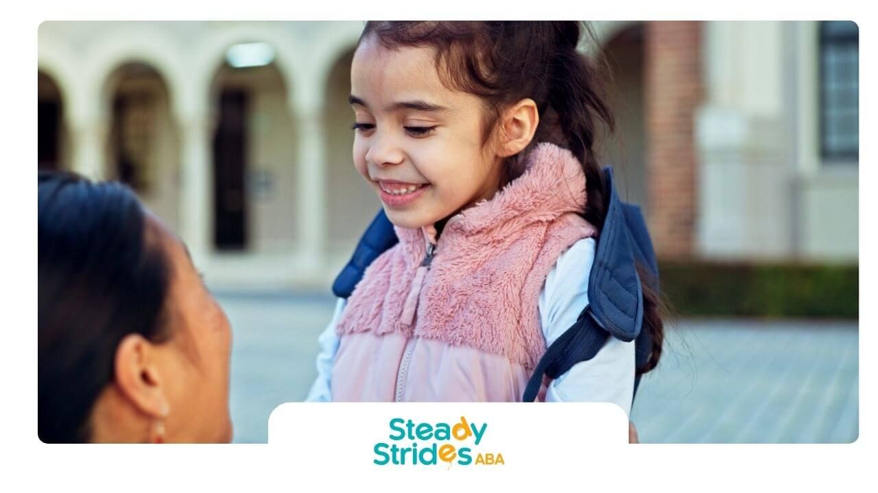 An autistic girl wearing backpack smiling at adult outside school building before or after class.