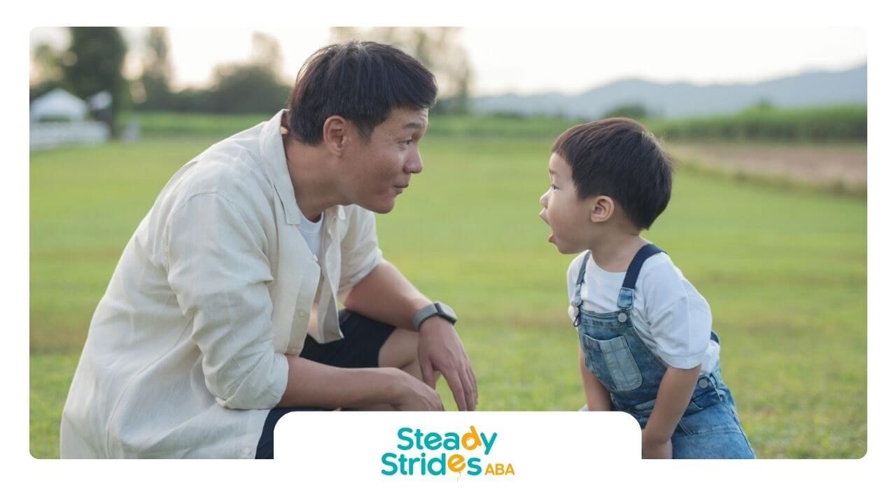 Male ABA therapist crouching and talking to a boy with autism outdoors on the grass.