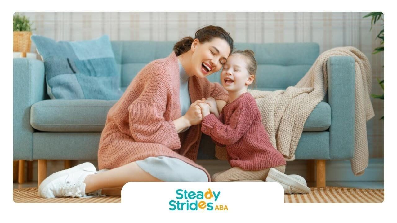 A BCBA and little girl with autism laughing together on the living room floor during ABA therapy.
