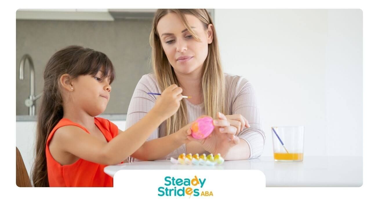 Therapist guiding autistic girl painting an egg at a table, encouraging creativity & learning.