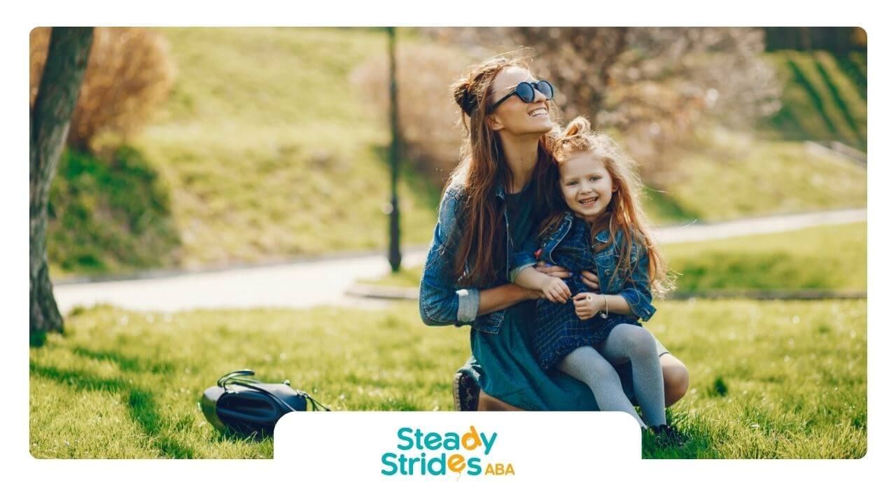 BCBA and young girl with autism smiling and hugging while sitting on grass in a sunny park.