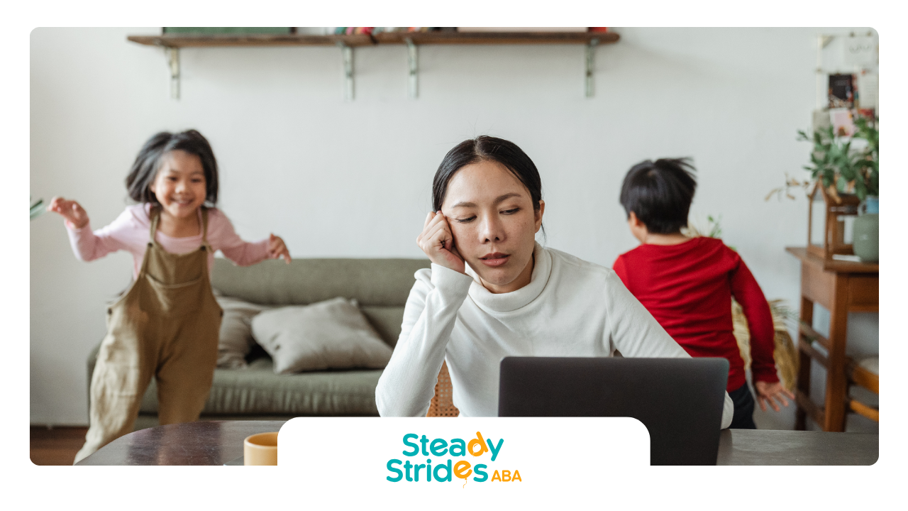 Woman at laptop, looking weary, with two children running around in living room.