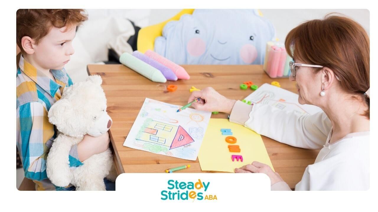 Therapist sits at a table guiding an autistic boy holding a teddy bear through drawing activity.