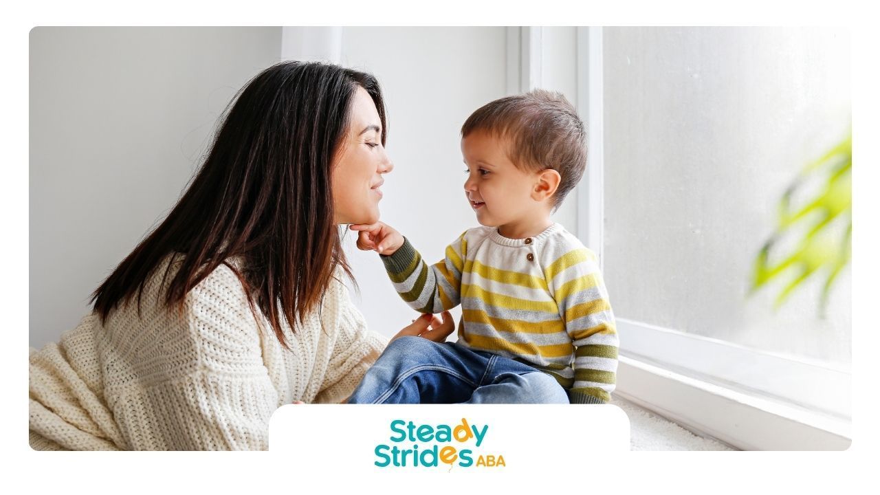 Woman and toddler look at each other lovingly near a window, child touches woman's chin.