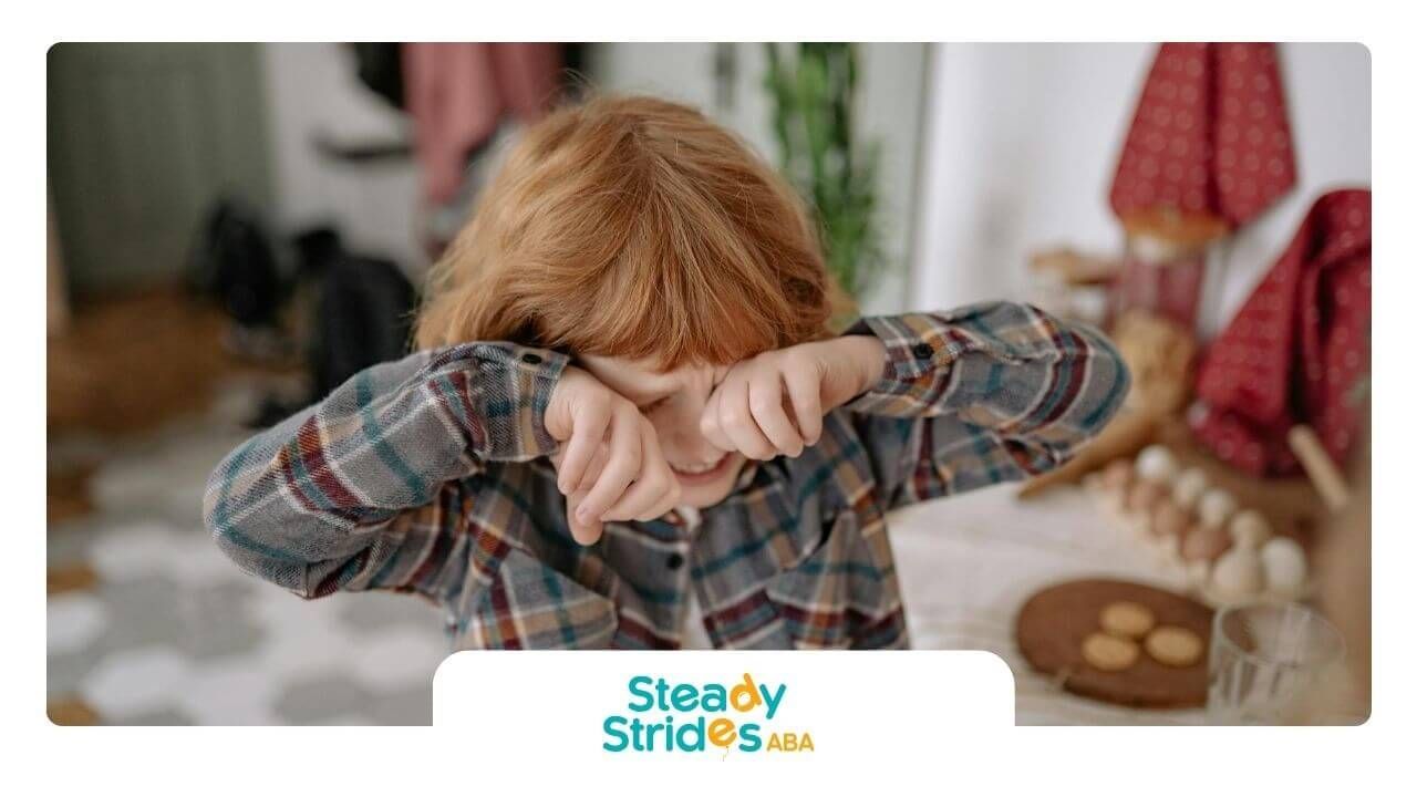 Red-haired autistic child in plaid shirt covers face near table with cookies and baking items