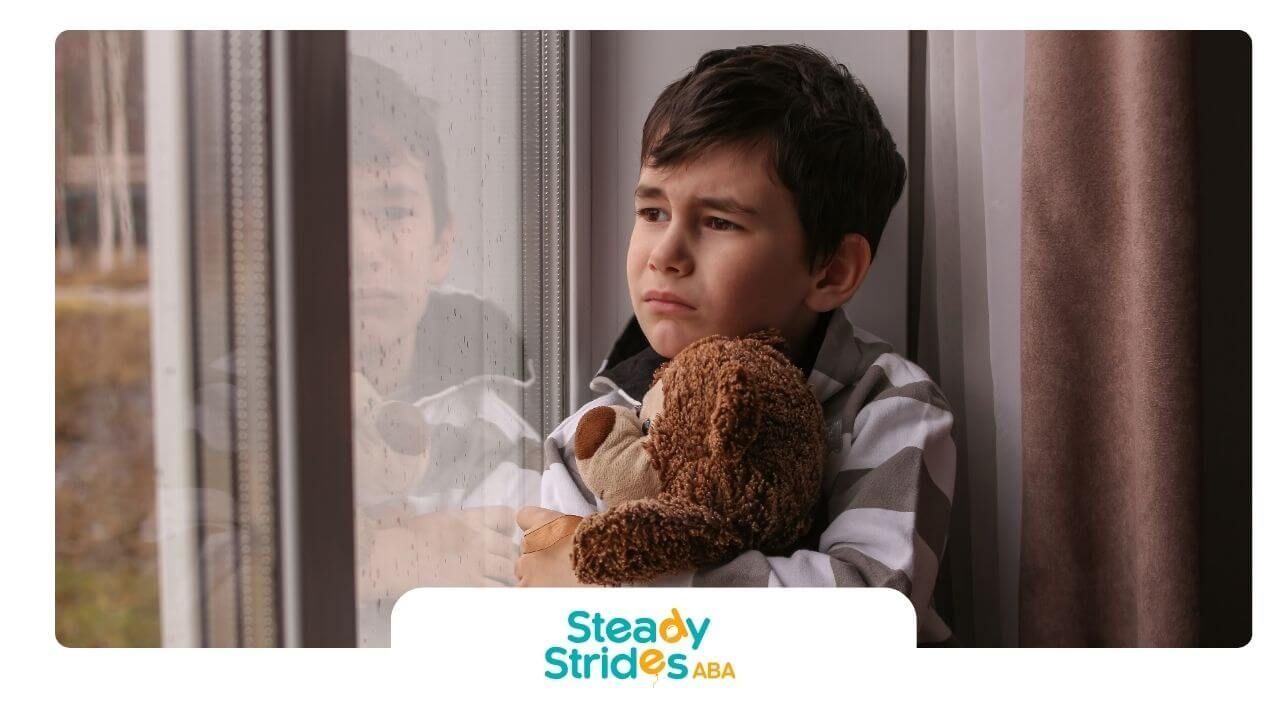 Sad autistic child hugs a teddy bear while looking out a rainy window, expressing loneliness.