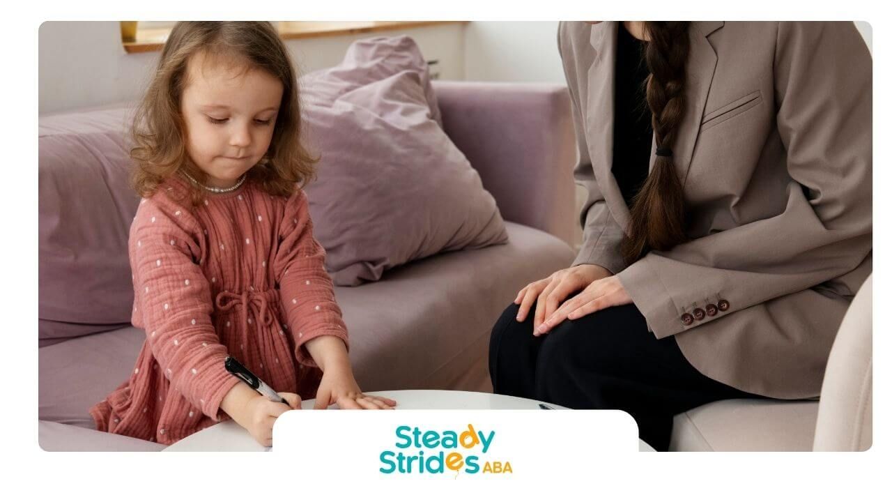 Autistic girl drawing on paper during therapy session while seated beside adult therapist on couch.