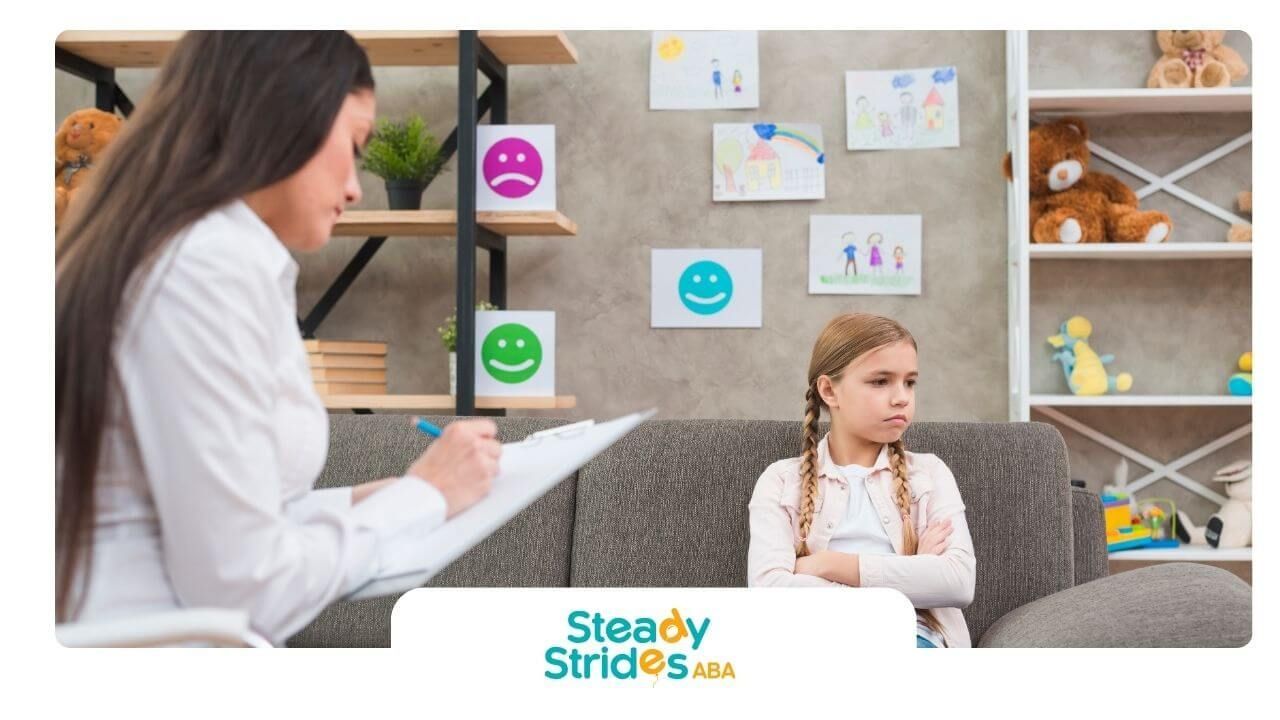 A therapist taking notes while autistic girl sits with arms crossed during child counseling session.
