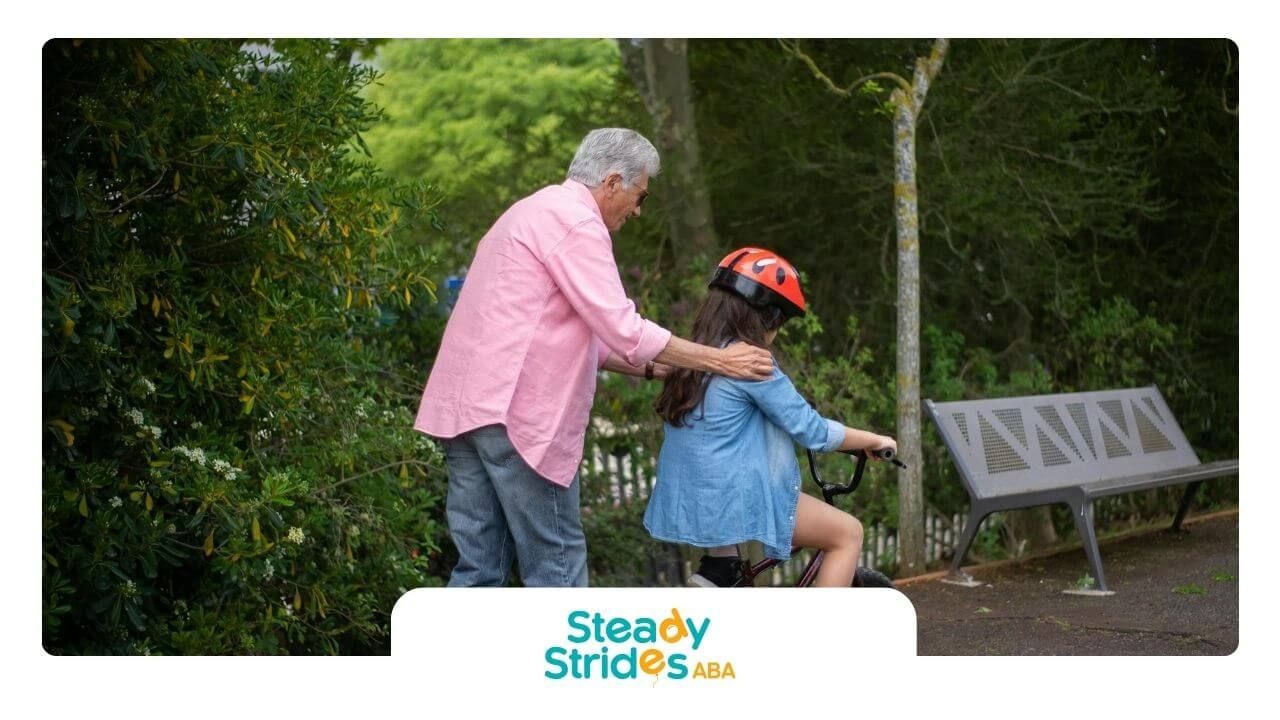 Male ABA therapist helps autistic girl ride bike on park path surrounded by trees and greenery