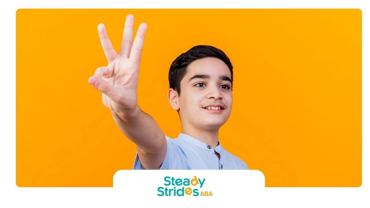 An autistic boy holds up three fingers against bright background, practicing number recognition.