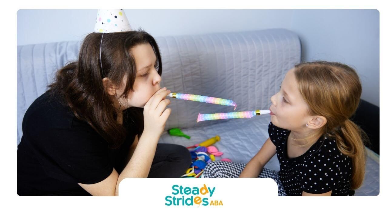 Two autistic girls wearing party hats blowing horns while sitting together during a celebration.