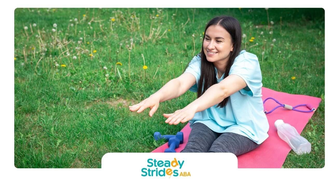 An autistic woman is doing yoga stretches on a pink mat outdoors in a grassy field with a smile.