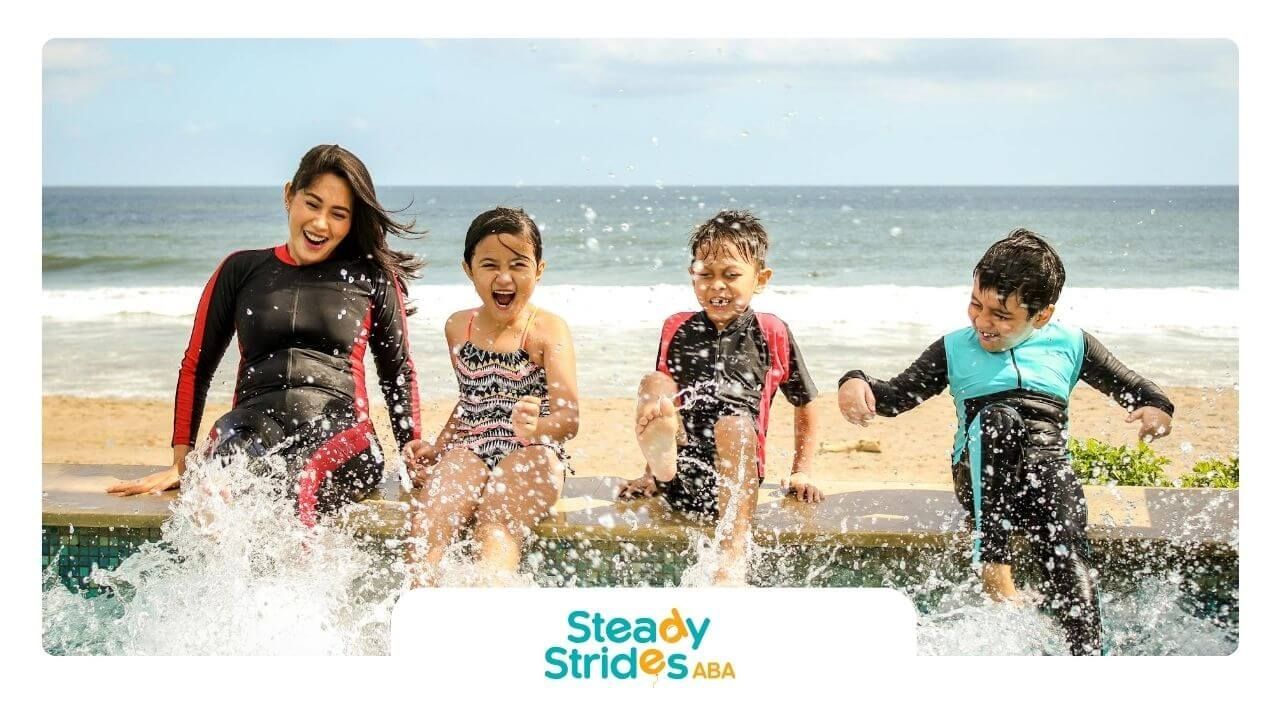 ABA therapist and three autistic kids splash feet in pool near beach under partly cloudy sky