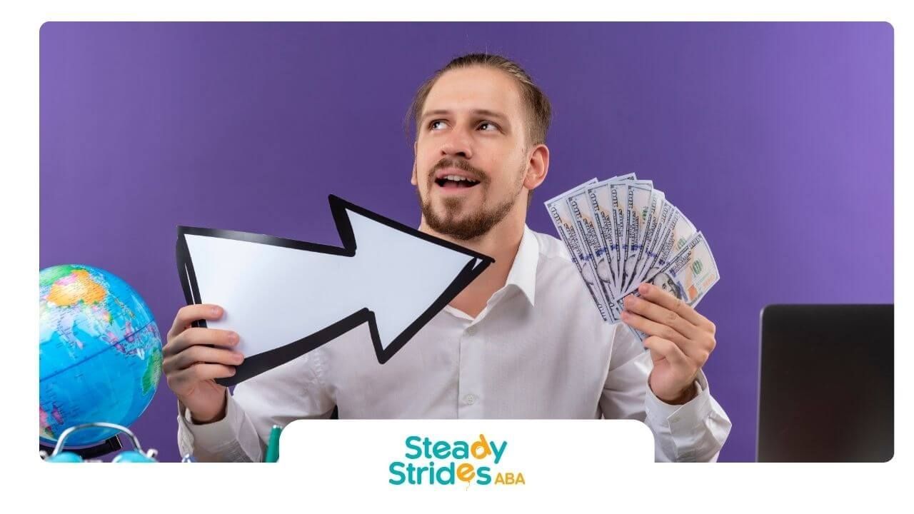 A male therapist holding cash and arrow sign at desk with globe, laptop, and travel items.