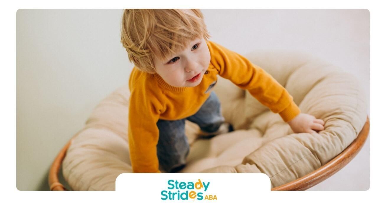 Autistic baby boy in yellow sweater climbing inside cushioned sensory chair at home during therapy.