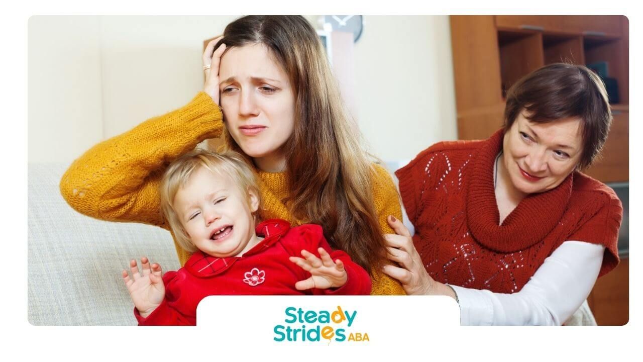 An ABA therapist holding crying autistic baby while older woman offers support in living room.
