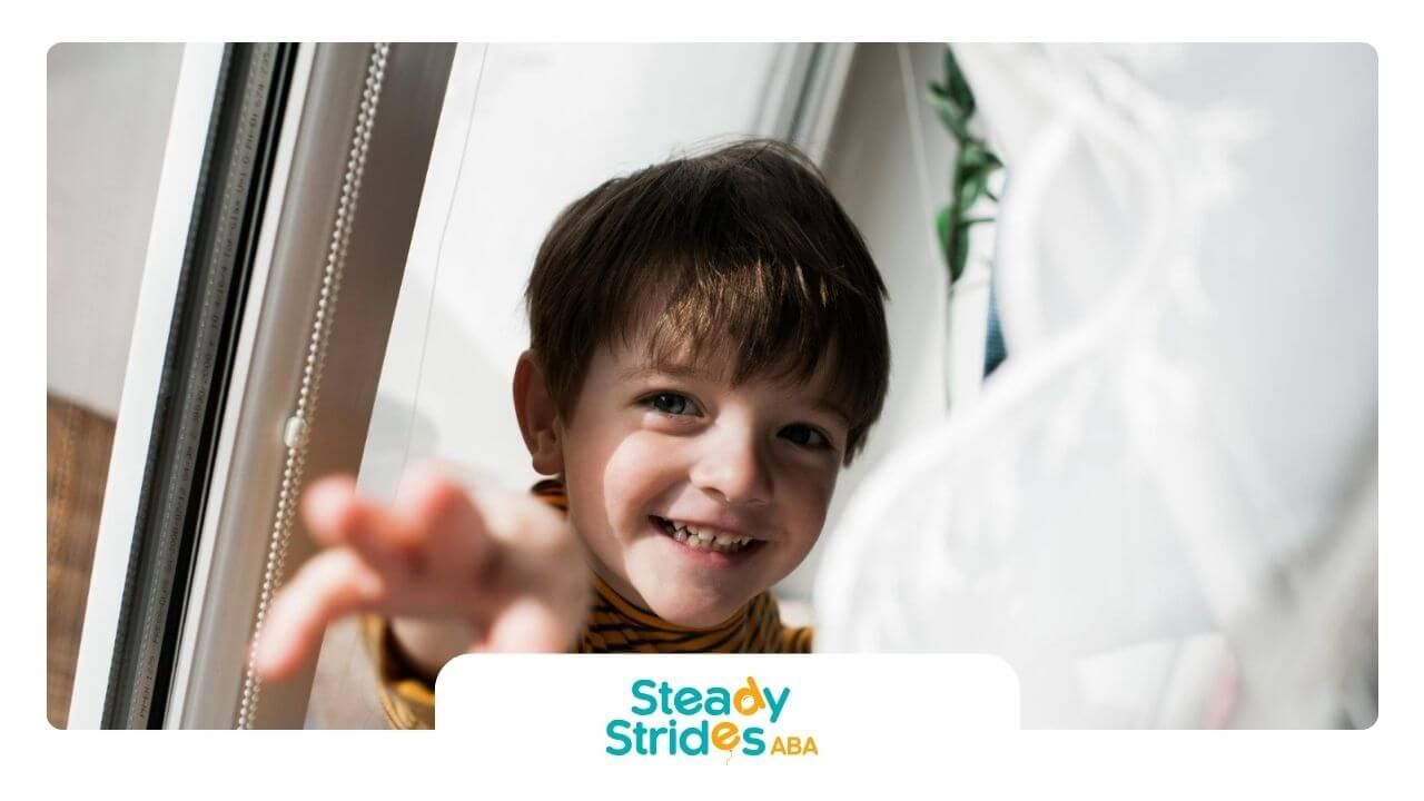A little boy with autism smiling and reaching toward the camera by a window.
