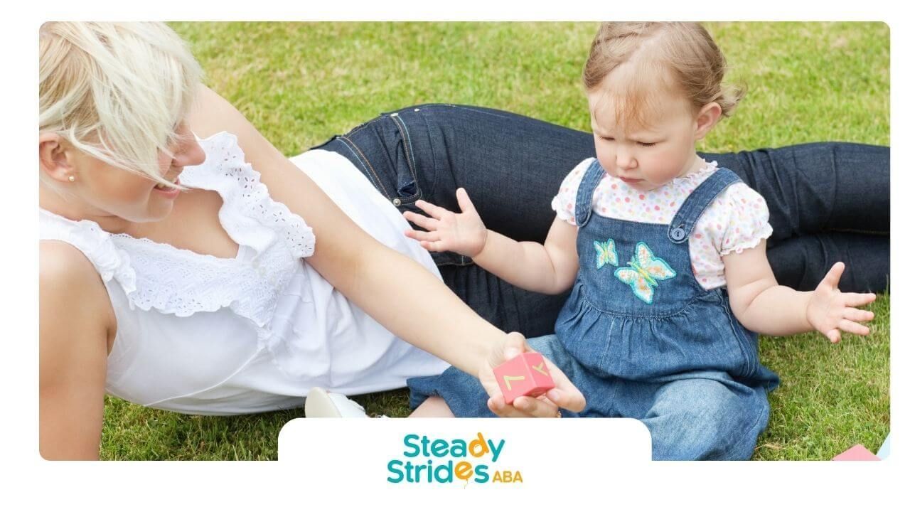 An ABA Therapist plays on grass with autistic toddler using colorful alphabet blocks outdoors.