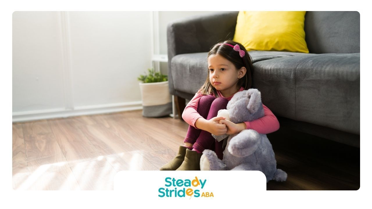 A young girl sits on the floor hugging a teddy bear, looking sad, near a sofa.