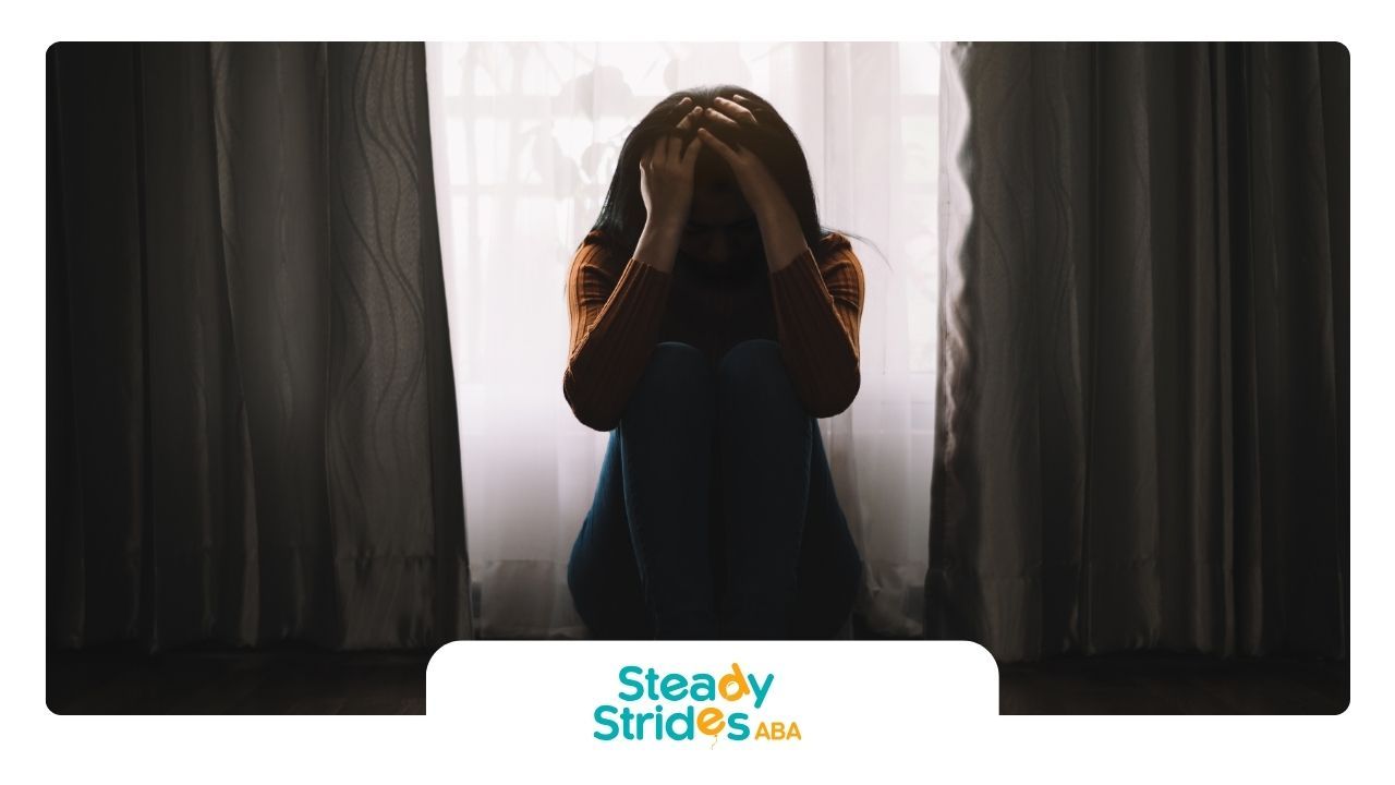 A woman is sitting in front of a window covering her face with her hands.