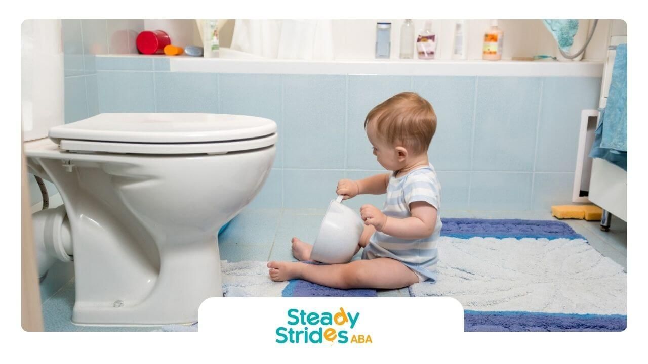 A toddler with autism holding a potty seat while sitting on the bathroom floor near a toilet.