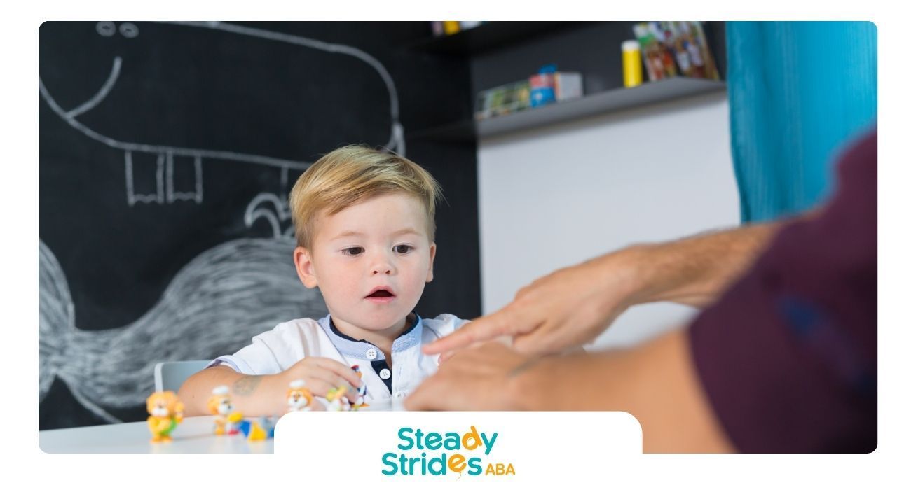 Boy with surprised expression at a table, playing with toys as an adult's hands interact. Black chalkboard wall in the background.