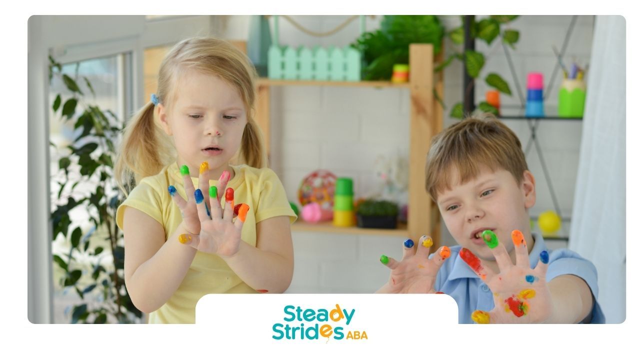 Two children, hands covered in colorful paint, looking surprised. Indoor setting.