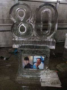 Clear ice sculpture reading “80” with two framed photos on a base, indoors at a celebration.