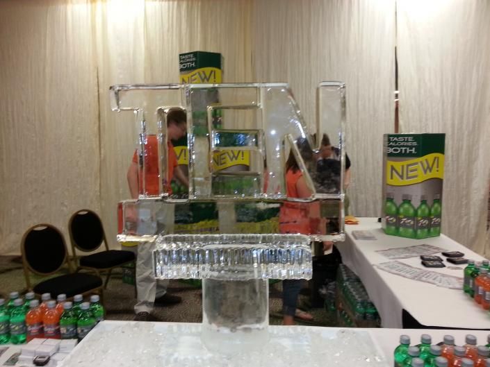 Trade show display with stacked green cans on a clear acrylic stand and a product table in the background