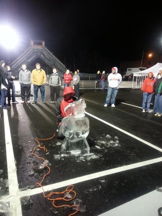 Child carving an ice block on a wet track at night, with several people watching nearby