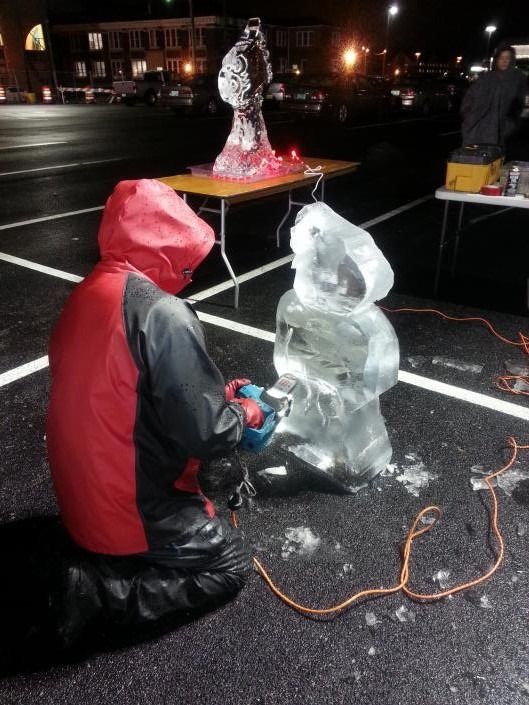 Person in red jacket kneels beside a clear ice sculpture at night in a wet parking lot.