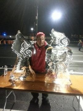 Person in a red jacket posing between two ice sculptures on a table at night.
