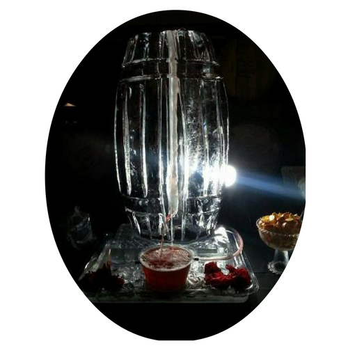Tall glass water fountain with bright light, on a dark table with red drinks and fruit bowls