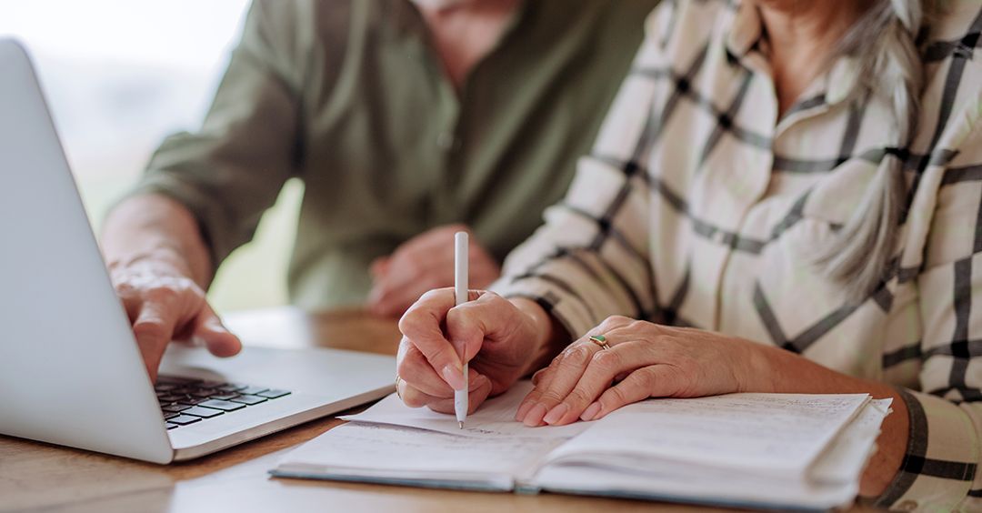 Two people working together at a table, one using a laptop and the other writing in a notebook.
