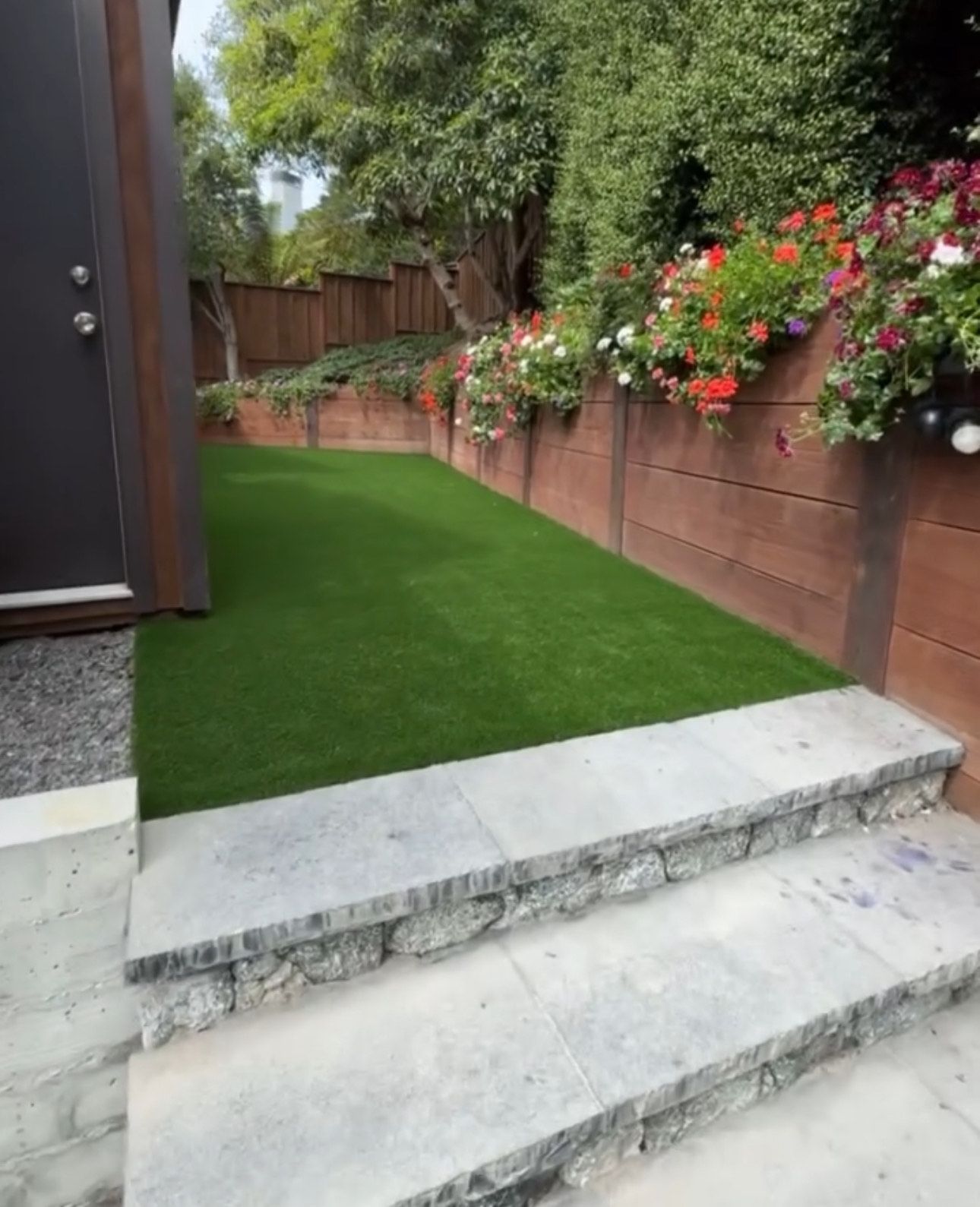 Stone steps lead to a small backyard with artificial green turf, bordered by a wooden wall with colorful flowers.