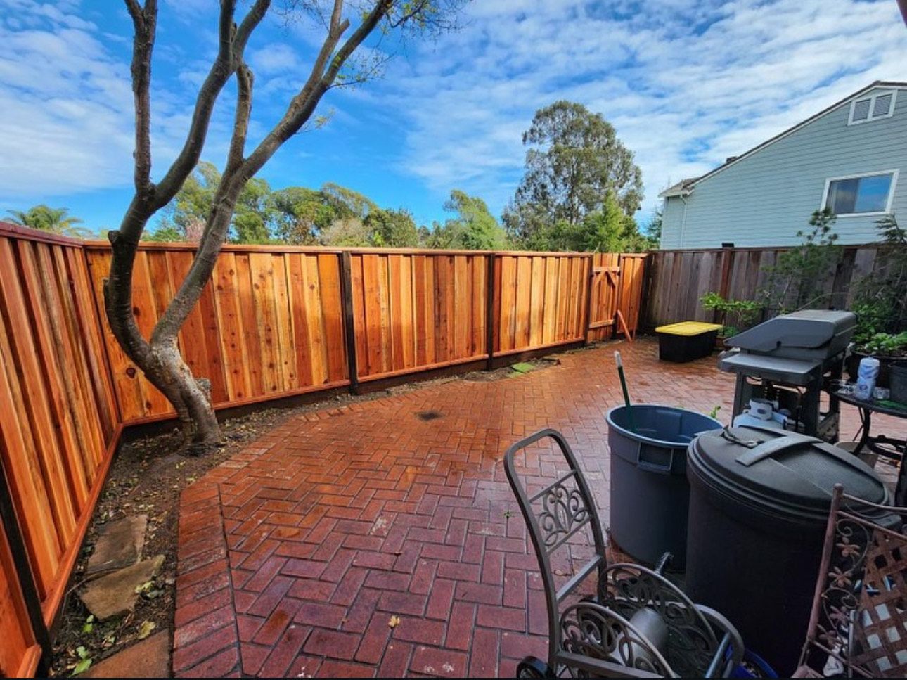 Red brick patio with wooden fence, tree, and grill under a blue sky.