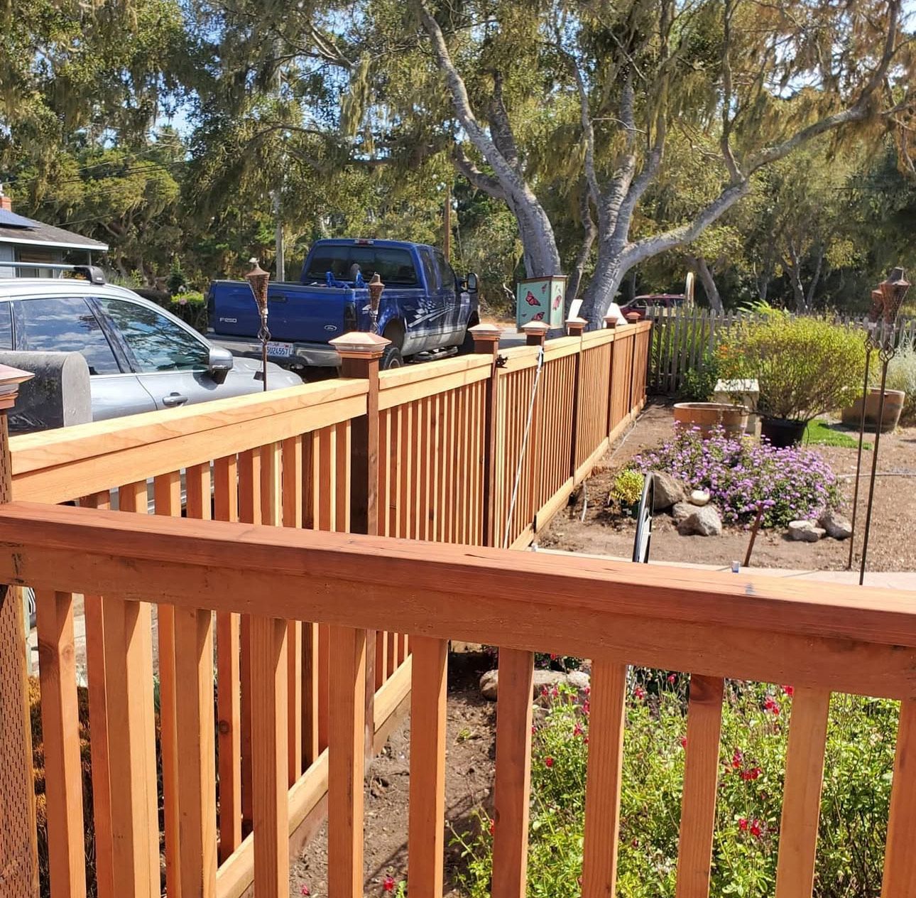 Wooden fence and railing, with a blue truck and car visible in the background. Sunny outdoor setting.