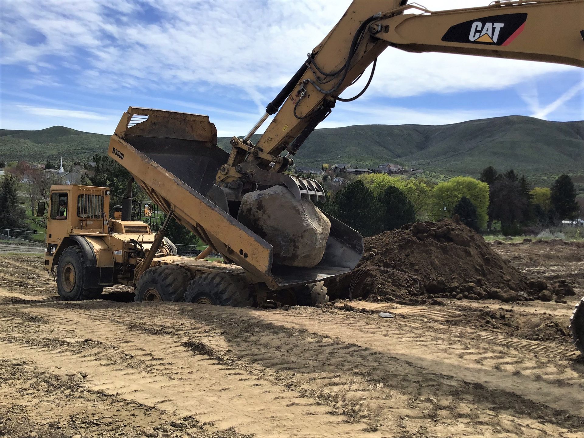 Excavator transferring a large rock. A yellow front-end loader is nearby on a dirt lot with a mountain backdrop.