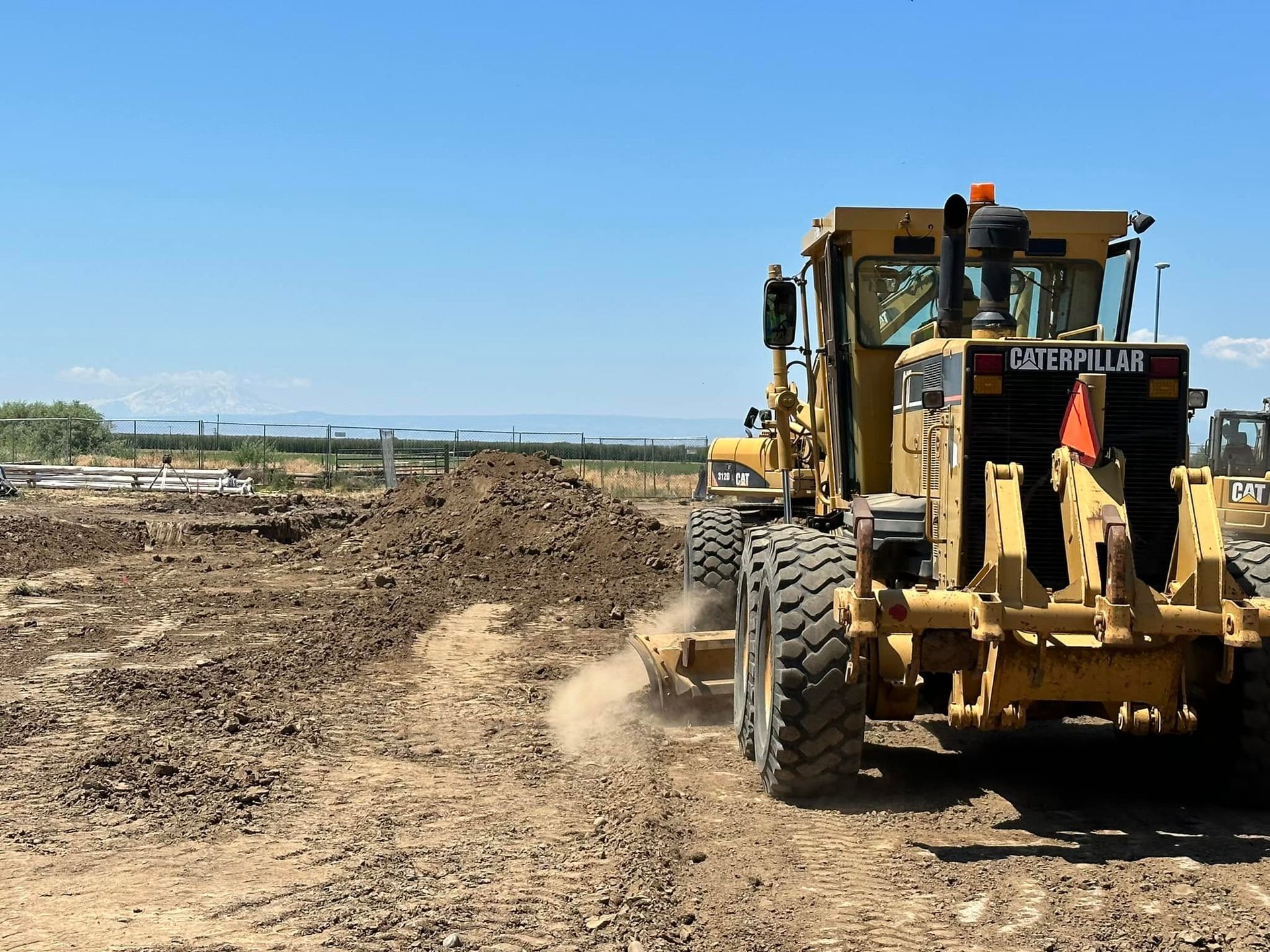 Yellow Caterpillar grader on a construction site, grading dirt under a clear blue sky.