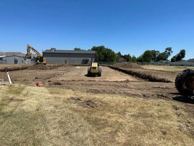 Construction site with heavy machinery excavating a rectangular area. A building and blue sky are in the background.