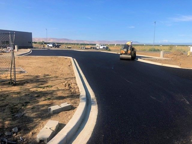 Asphalt paving with roller machine on a new road. Concrete curb, beige field, blue sky, and construction in the background.