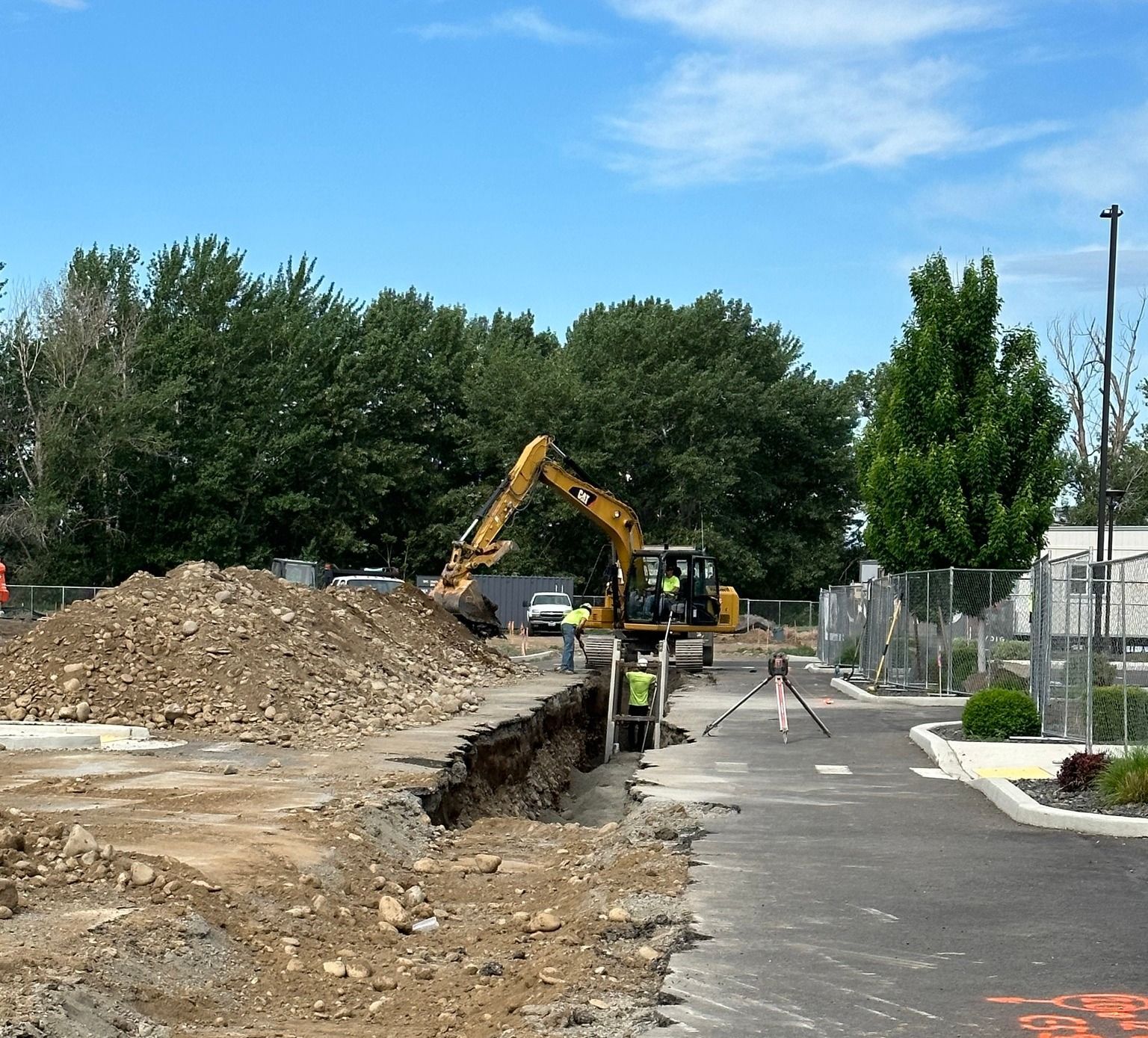 Excavator digging trench at construction site, dirt pile, workers, trees, blue sky.