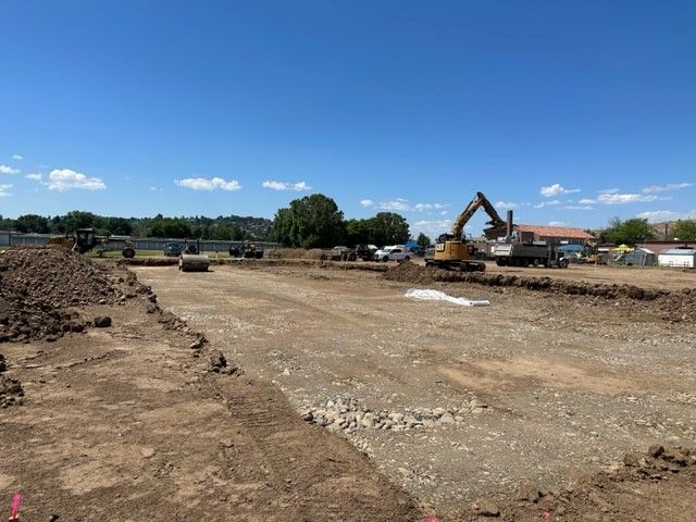 Construction site; earthmovers, compactors working on leveled ground, piles of dirt, blue sky.