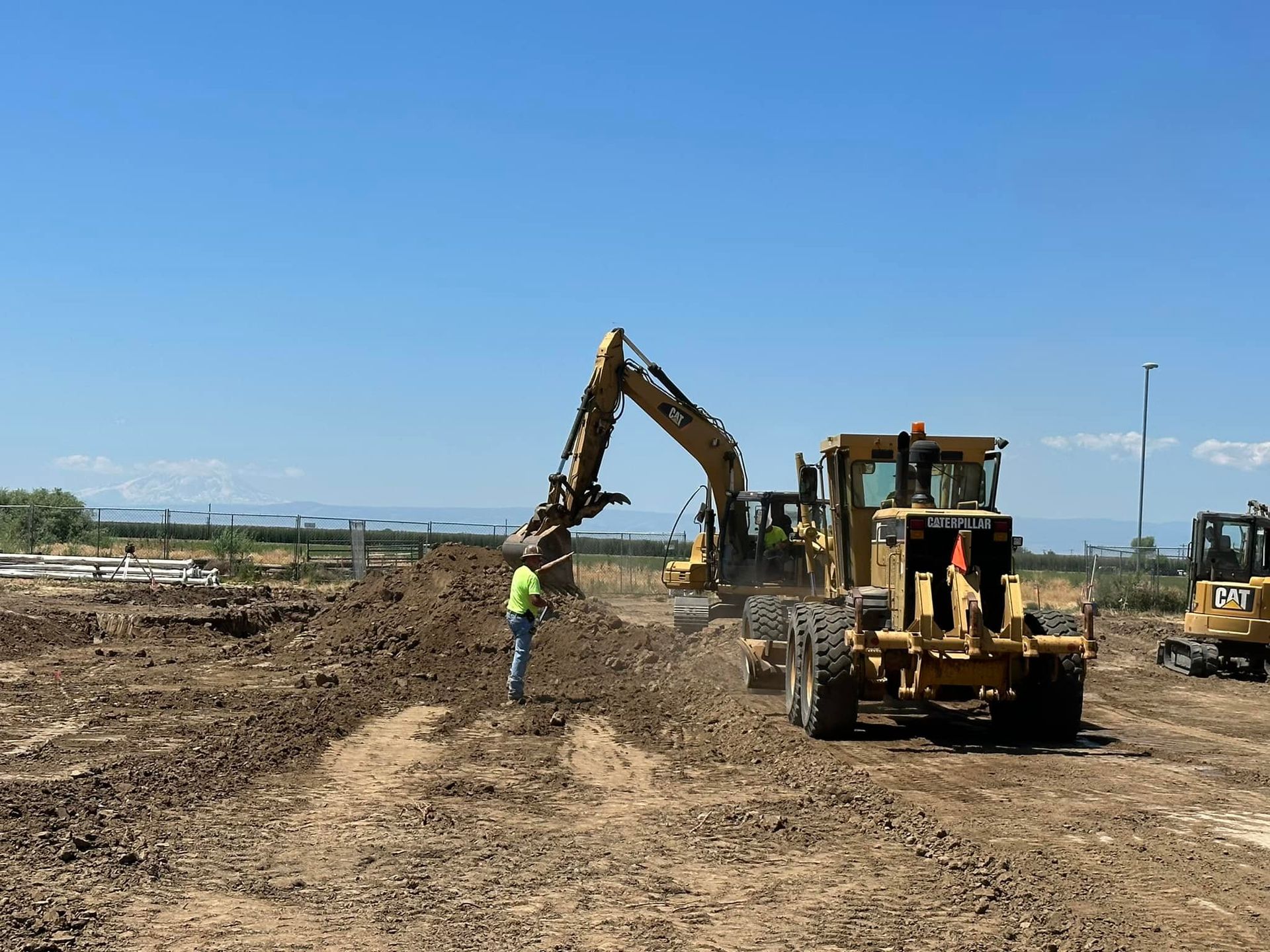 Construction site with excavator and grader moving dirt under a clear blue sky. A worker is observing.