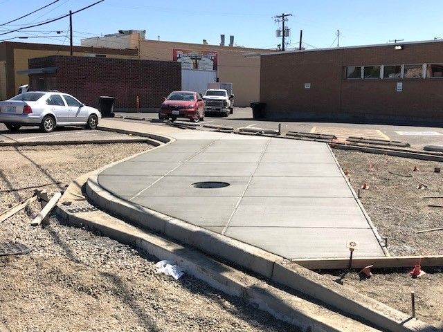 Newly poured concrete walkway with cars parked in the background and a building.