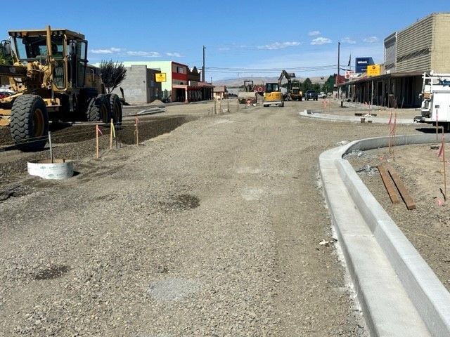 Street construction in progress; gravel roadbed, new curb, construction equipment, businesses in the background.