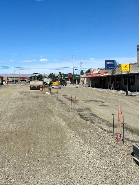 Construction on a town street. Heavy machinery, gravel road, storefronts, and a blue sky.